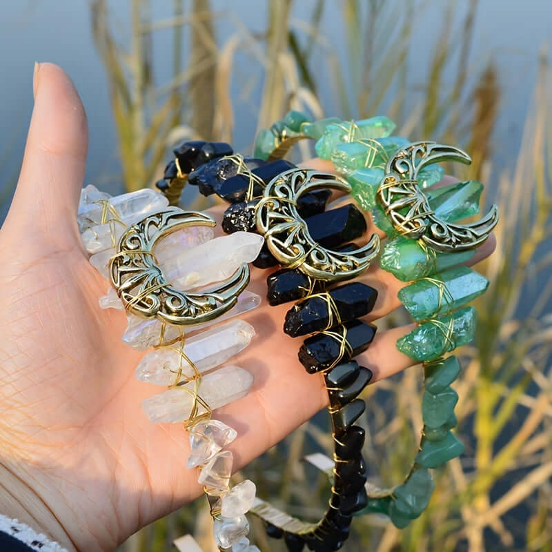 Hand holding three moon-shaped crystal headbands in white, black, and green, with natural crystals and boho-chic design.