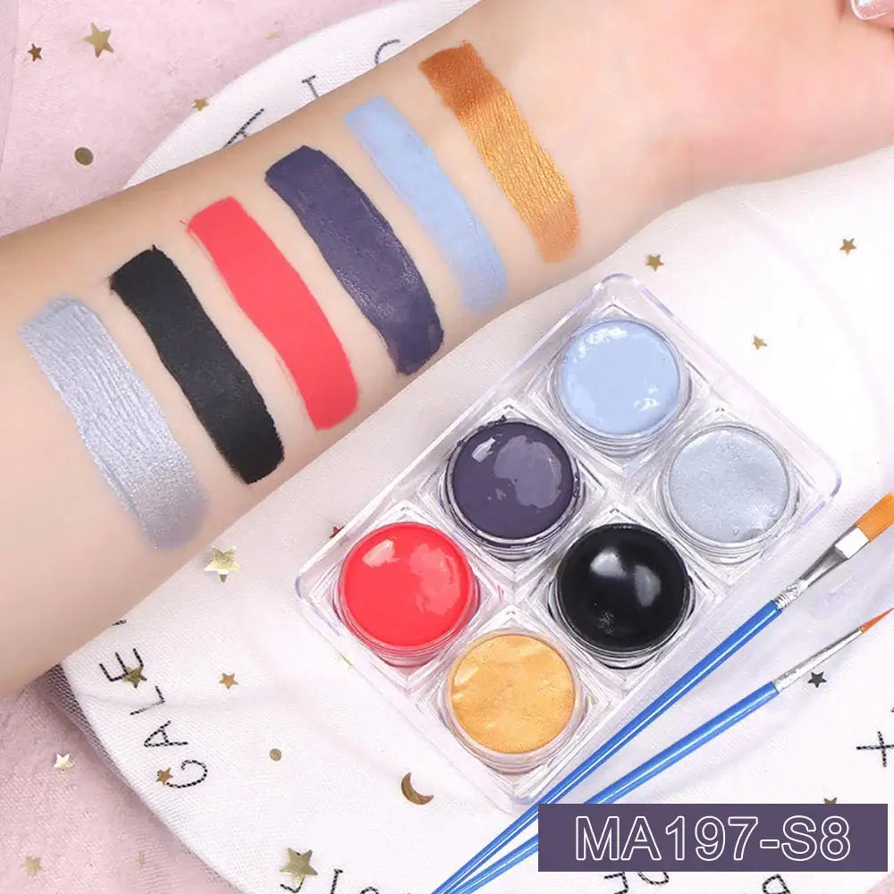 Makeup swatches on arm with 6 bold colors, silver, black, red, blue, purple, and gold, beside open glitter gel jars and brushes.