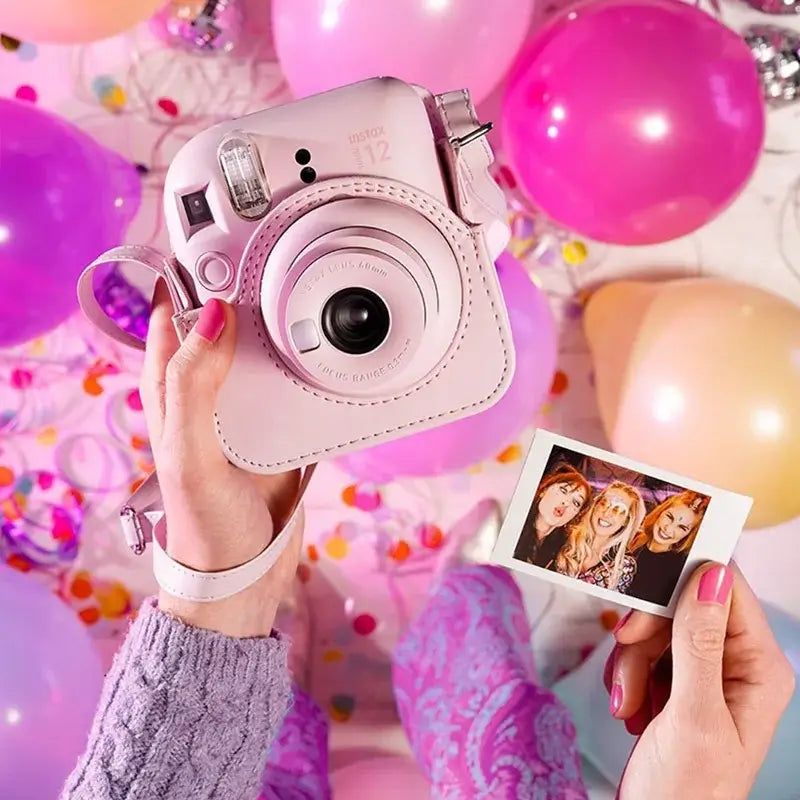 Person holding Fujifilm Instax Mini 12 Instant Camera in Blossom Pink, surrounded by colorful balloons, with an instant photo.