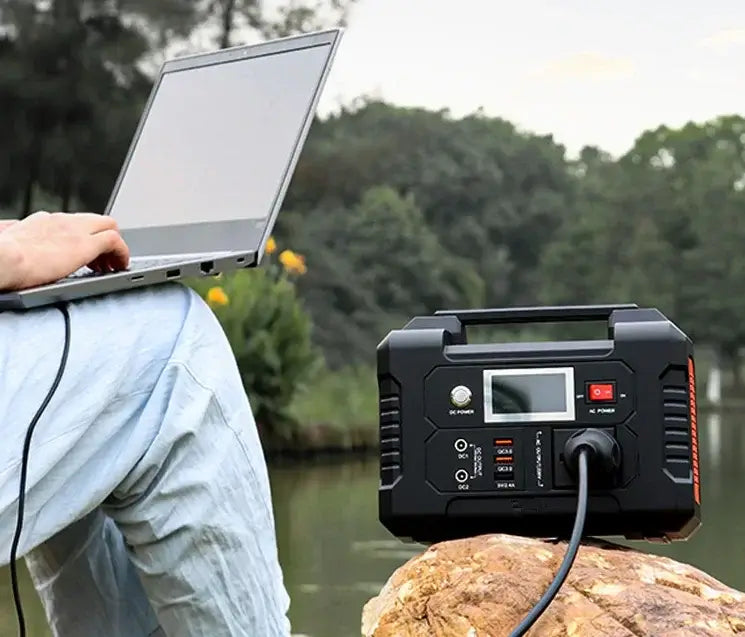 Portable power station charging a laptop outdoors near a lake with trees in the background.