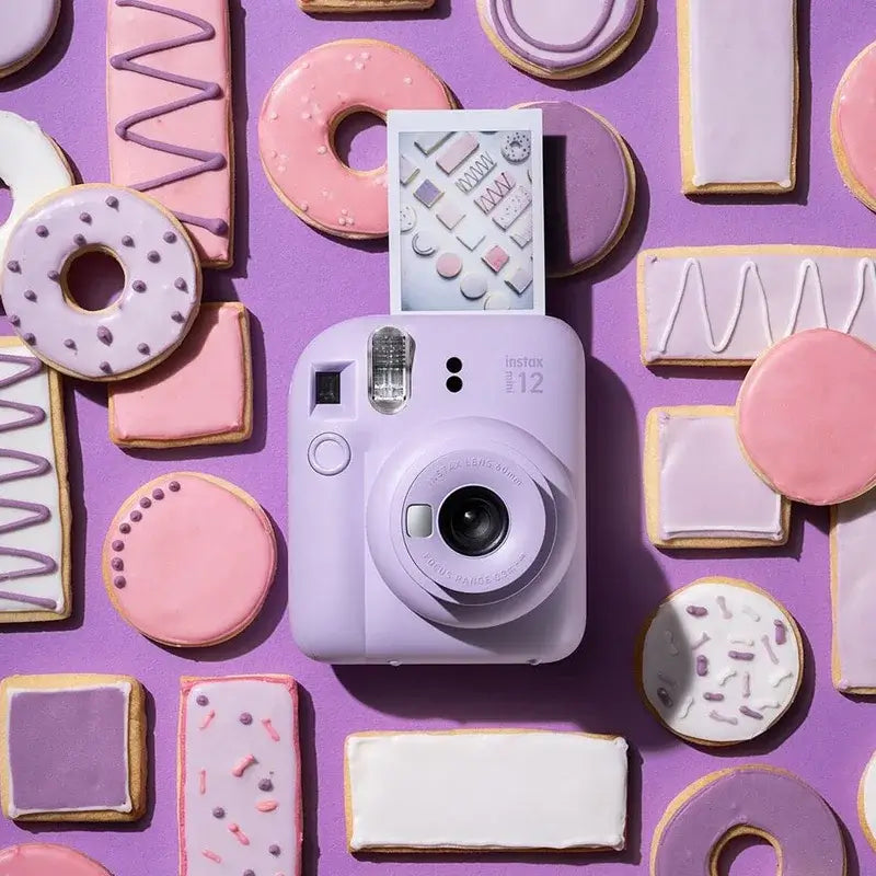 Lilac Fujifilm Instax Mini 12 instant camera surrounded by trendy purple and pink donuts and cookies, capturing stylish photography.