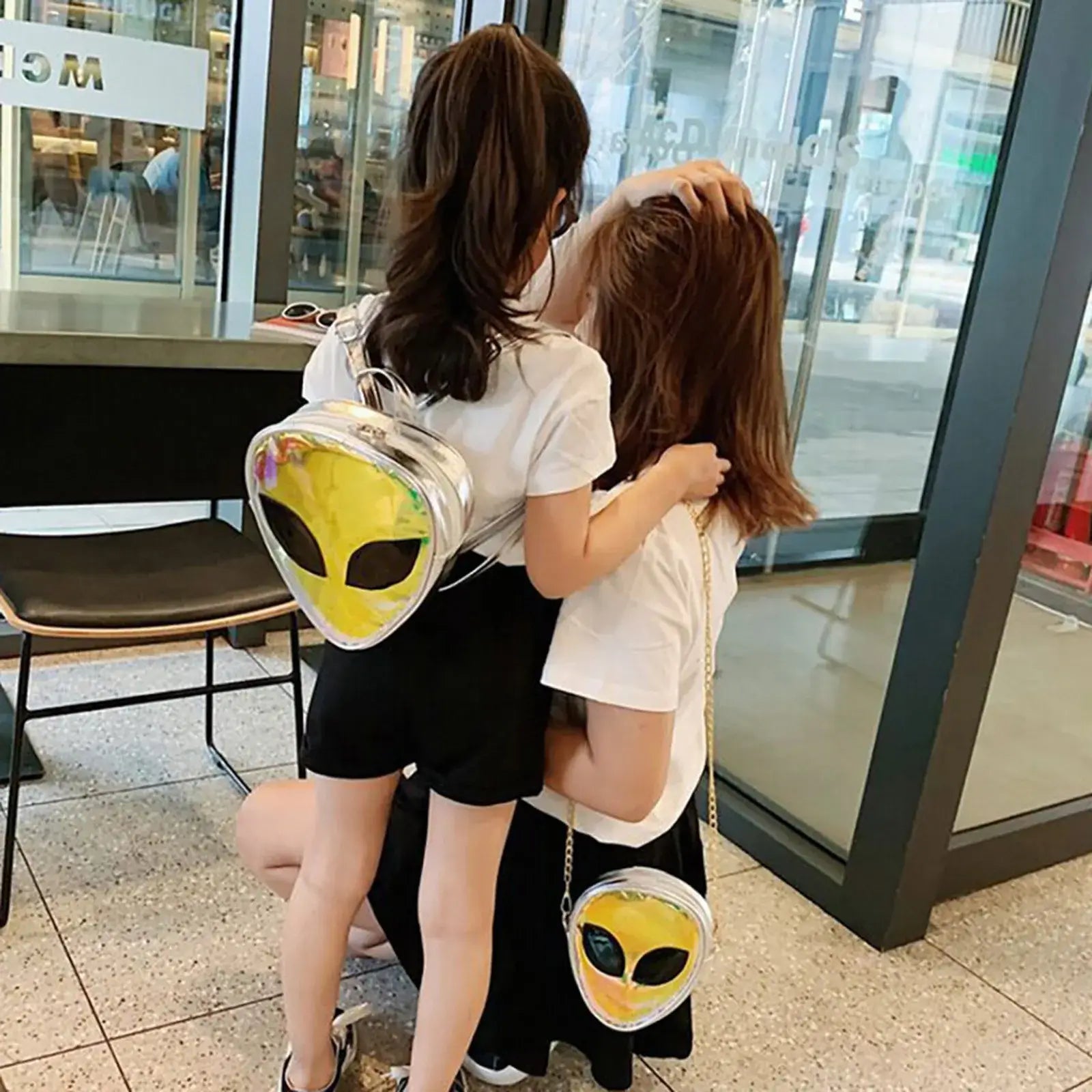 Mother and daughter wearing transparent jelly alien shoulder bags in a café, showcasing trendy festival fashion backpacks.