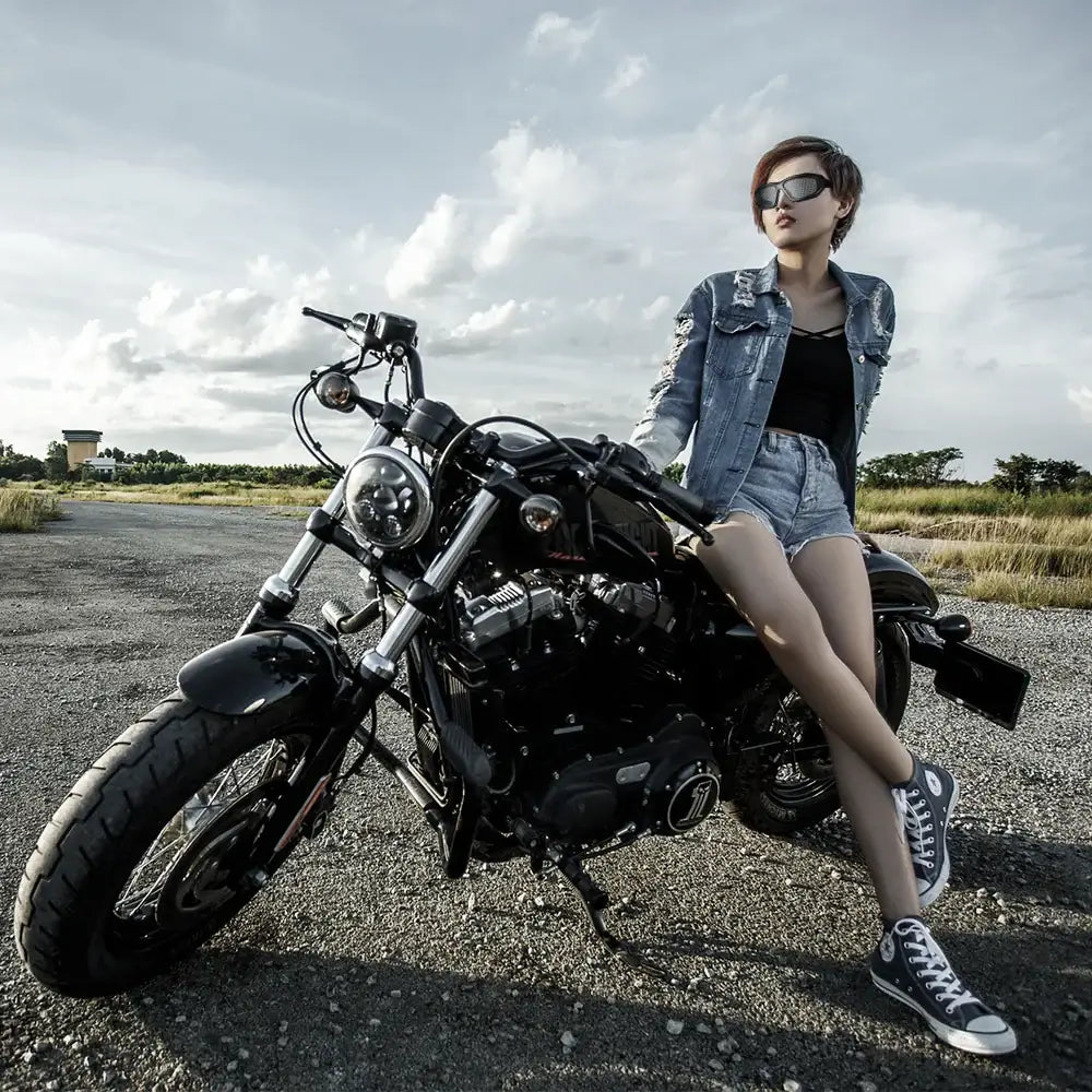 Person wearing sunglasses and denim jacket sitting on a black motorcycle in an open field.