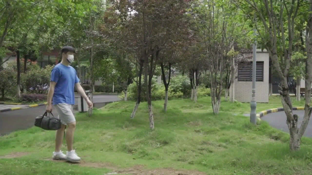 Person walking through a park wearing a mask and carrying a black bag, surrounded by greenery and trees.